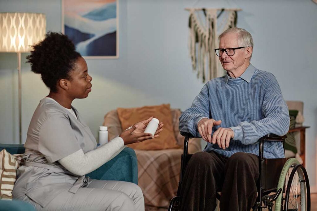 An older adult chats with a friend in a cozy living room surrounded by personal belongings and a small dog.